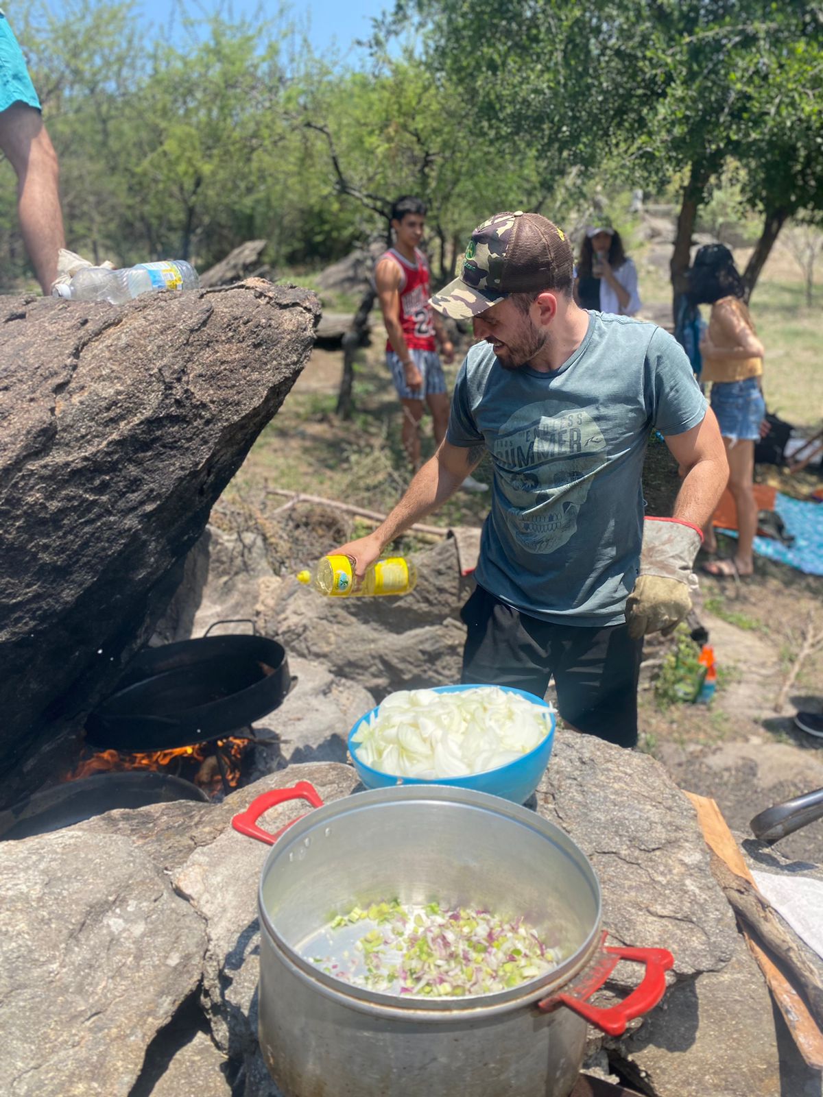 Ecolink. Un hombre cocinando al aire libre, junto a ollas llenas de ingredientes frescos como cebolla y pimiento, en una zona rocosa rodeada de árboles. Representa convivencia, cocina comunitaria y experiencias en la naturaleza.