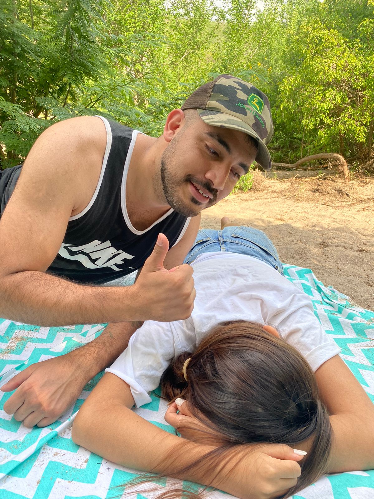 Ecolink. Un hombre sonriente, con gorra camuflada, haciendo un gesto de aprobación con el pulgar hacia arriba mientras una mujer descansa sobre una manta en el suelo, en un entorno natural. Transmite un momento de descanso.